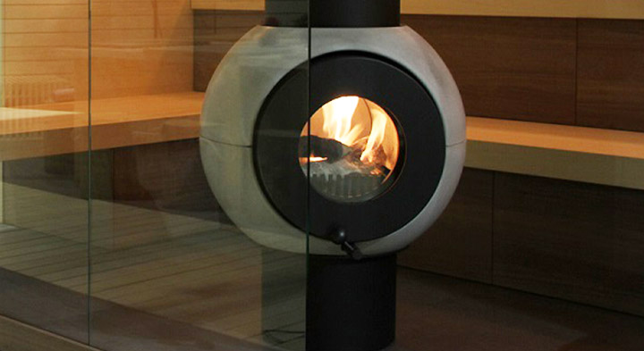 Grau-schwarzer firetube Ofen mit sichtbarem Feuer in einer Sauna, umgeben von Glas und Holzsitzbänken.