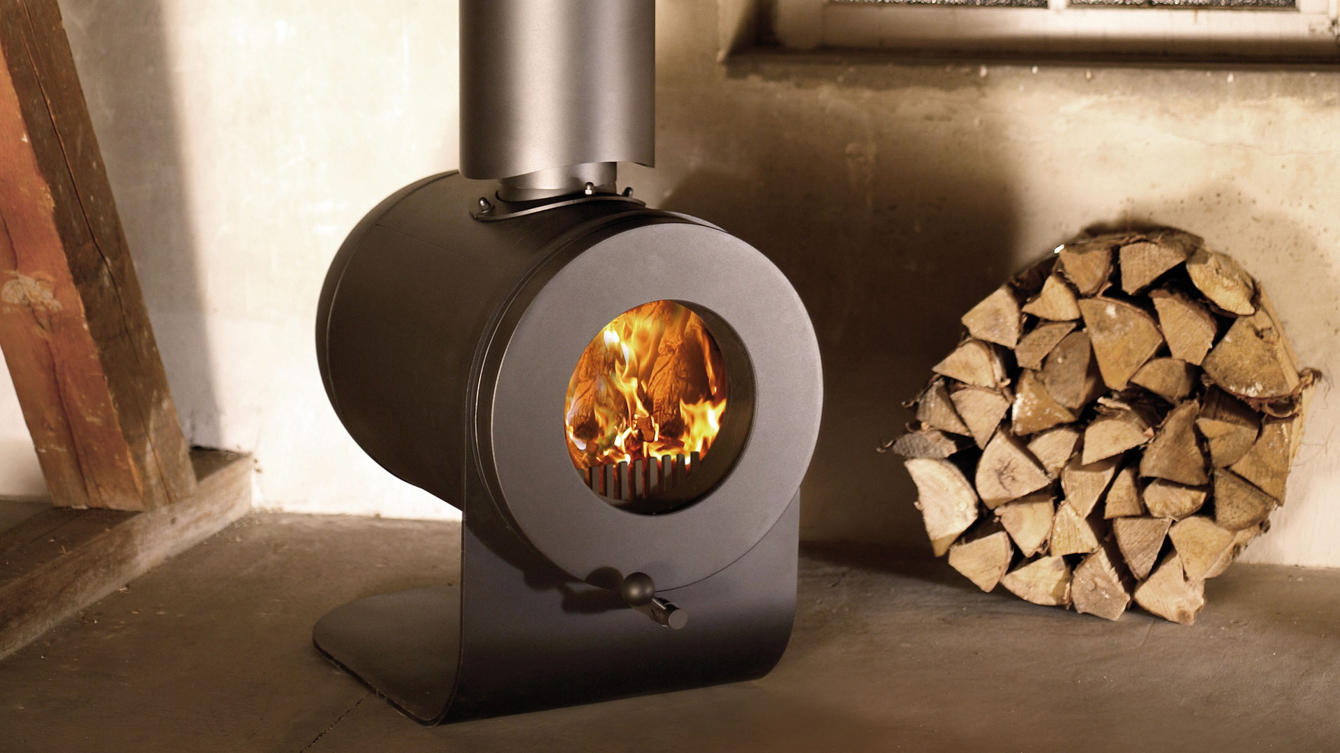 Auf dem Boden stehender firetube Speicherofen mit sichtbarem Feuer, in rustikalem Raum; daneben Holzscheite gestapelt.