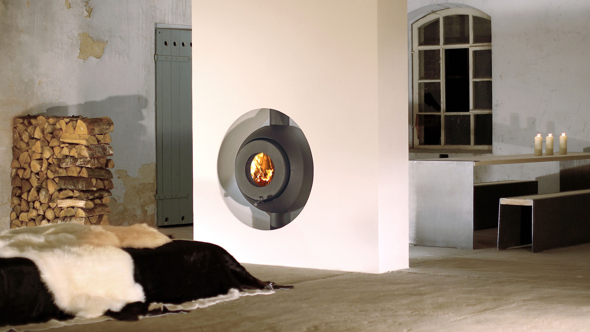 Schwarzer firetube Ofen mit sichtbarem Feuer, eingebaut in raumstehende Wand; dahinter Tisch und Holzscheite.