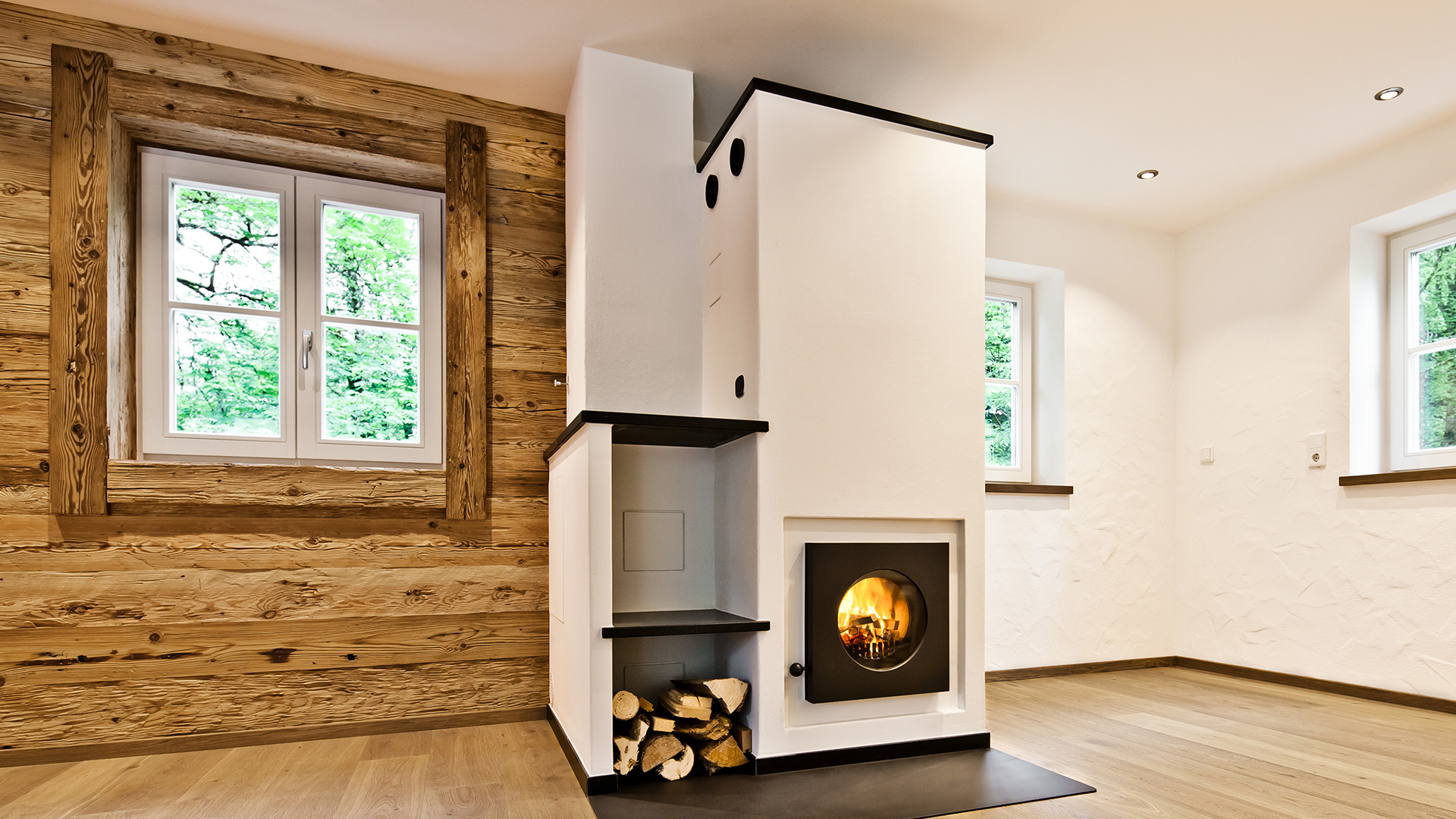 Firetube Ofen mit sichtbarem Feuer in einer trennenden Wand integriert; daneben Fach mit Holzscheiten, alles weiß verkleidet; linke Raumseite Holzwand, rechte Seite weiße Betonwand.