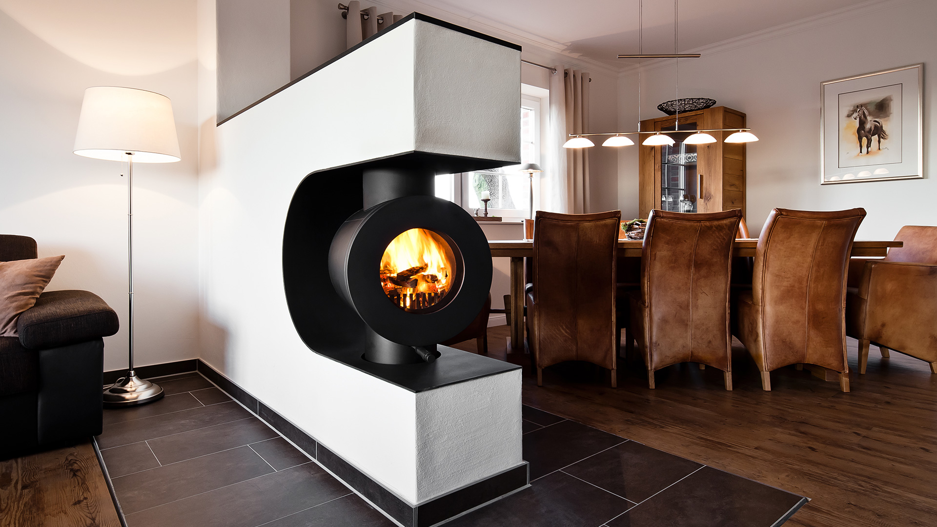 Schwarzer firetube Ofen mit sichtbarem Feuer, in eine raumtrennende Wand integriert; im Hintergrund ein Wohnzimmer.
