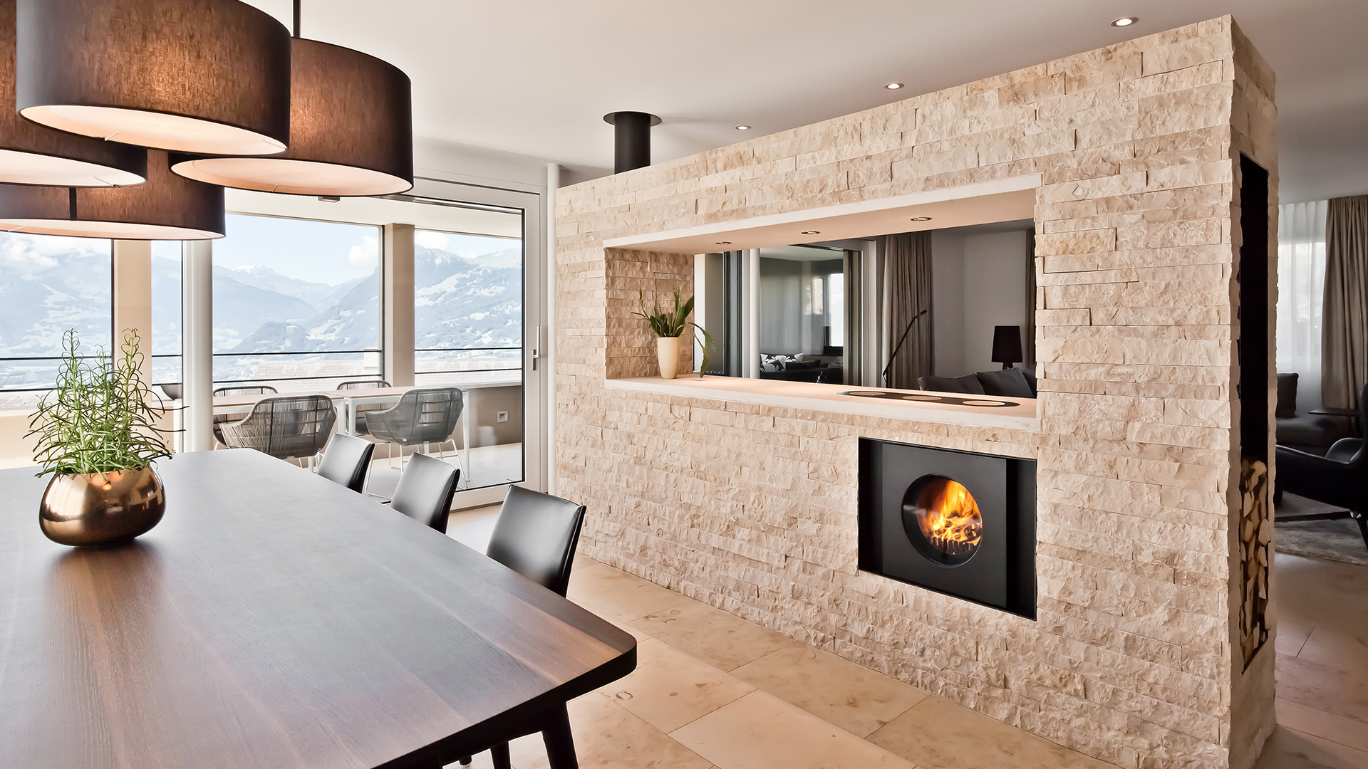 Wohnzimmer mit Esstisch und Fensterfront mit Aussicht; steinverkleidete Trennwand mit integriertem schwarzem firetube Ofen und Kochplatten mit Küchen und sichtbarem Feuer.