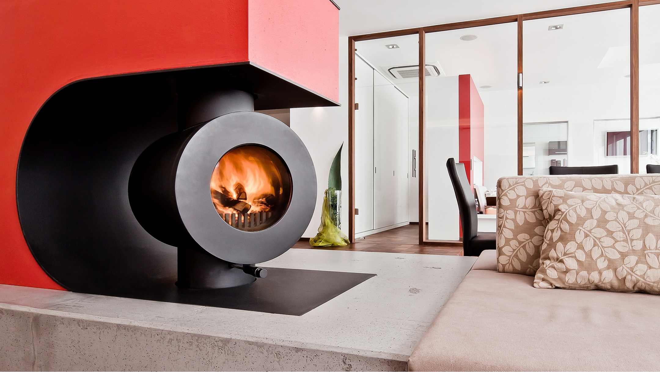 In eine Wohnlandschaft integrierter schwarzer firetube Speicherofen mit sichtbarem Feuer in rotem Wandausschnitt mit Sitzfläche; im Hintergrund ein Raum mit Glaswänden.