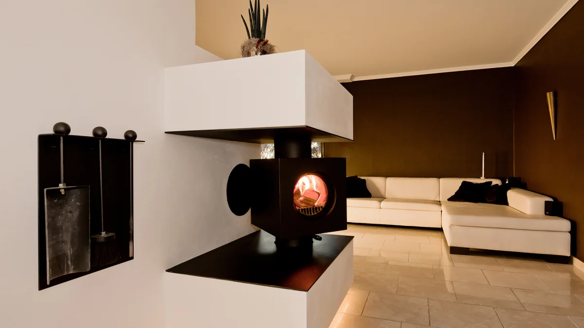 Schwarzer firetube Ofen mit sichtbarem Feuer, in eine raumtrennende Wand integriert; im Hintergrund ein Wohnzimmer.
