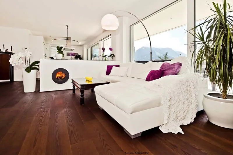 Weißes Sofa in einem Wohnzimmer; links davon trennende Wand mit integriertem schwarzem firetube Ofen und sichtbarem Feuer, im Hintergrund Fenster mit Ausblick.