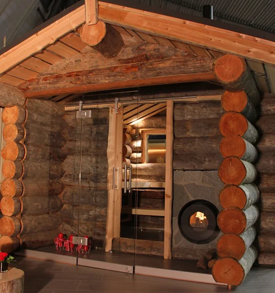 Holzhütte von außen mit Vorraum aus Glaswand und Glastür; darin ein firetube Ofen mit sichtbarem Feuer, in die Hauswand integriert.
