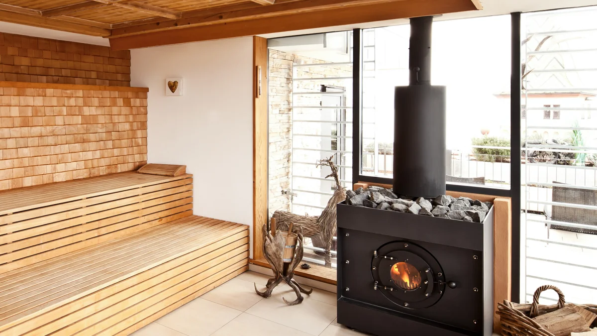 Firetube Saunaofen in einer Sauna mit Glaswand; daneben Holzdekoration, Korb mit Holzscheiten auf dem Boden und Sitzbänke aus Holz.