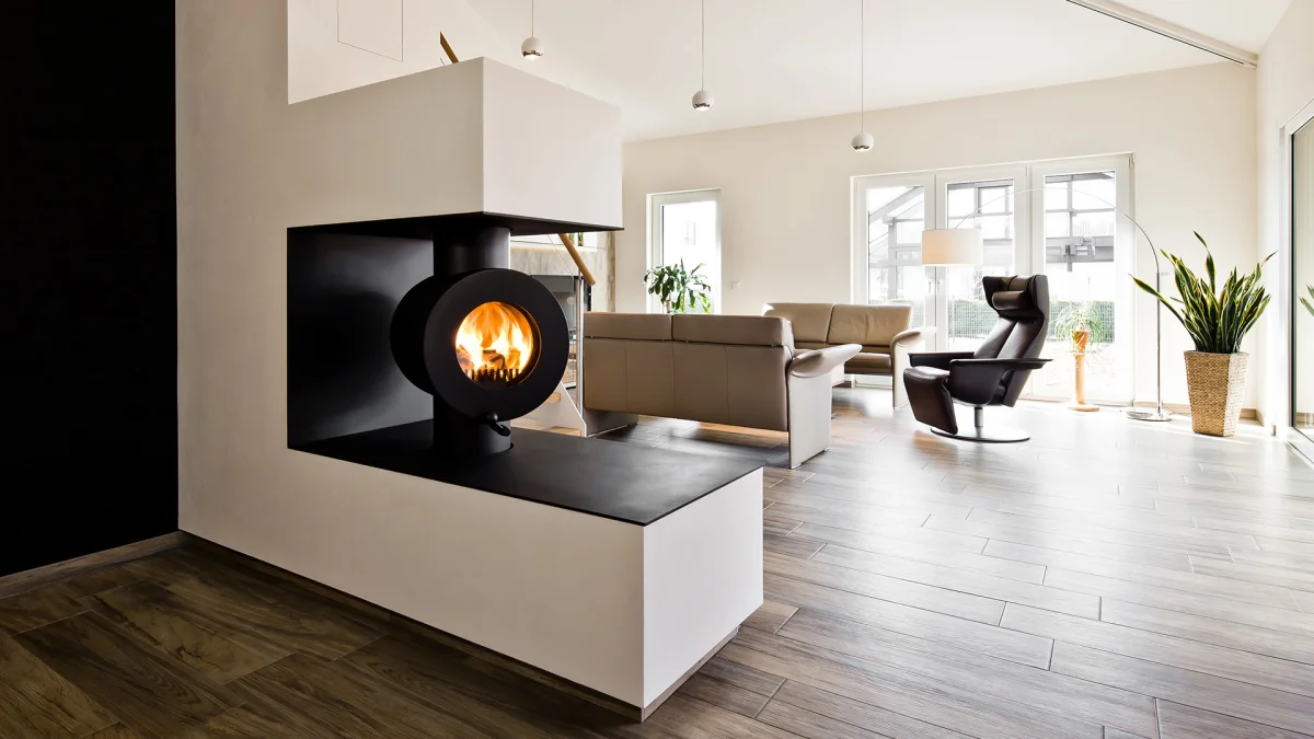 Schwarzer firetube Ofen mit sichtbarem Feuer, in eine raumtrennende Wand integriert; im Hintergrund ein Wohnzimmer.