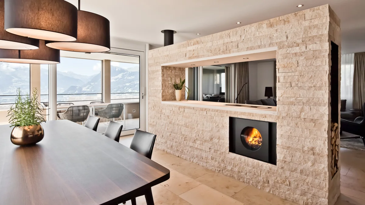 Wohnzimmer mit Esstisch und Fensterfront mit Aussicht; steinverkleidete Trennwand mit integriertem schwarzem firetube Ofen und Kochplatten mit Küchen und sichtbarem Feuer.