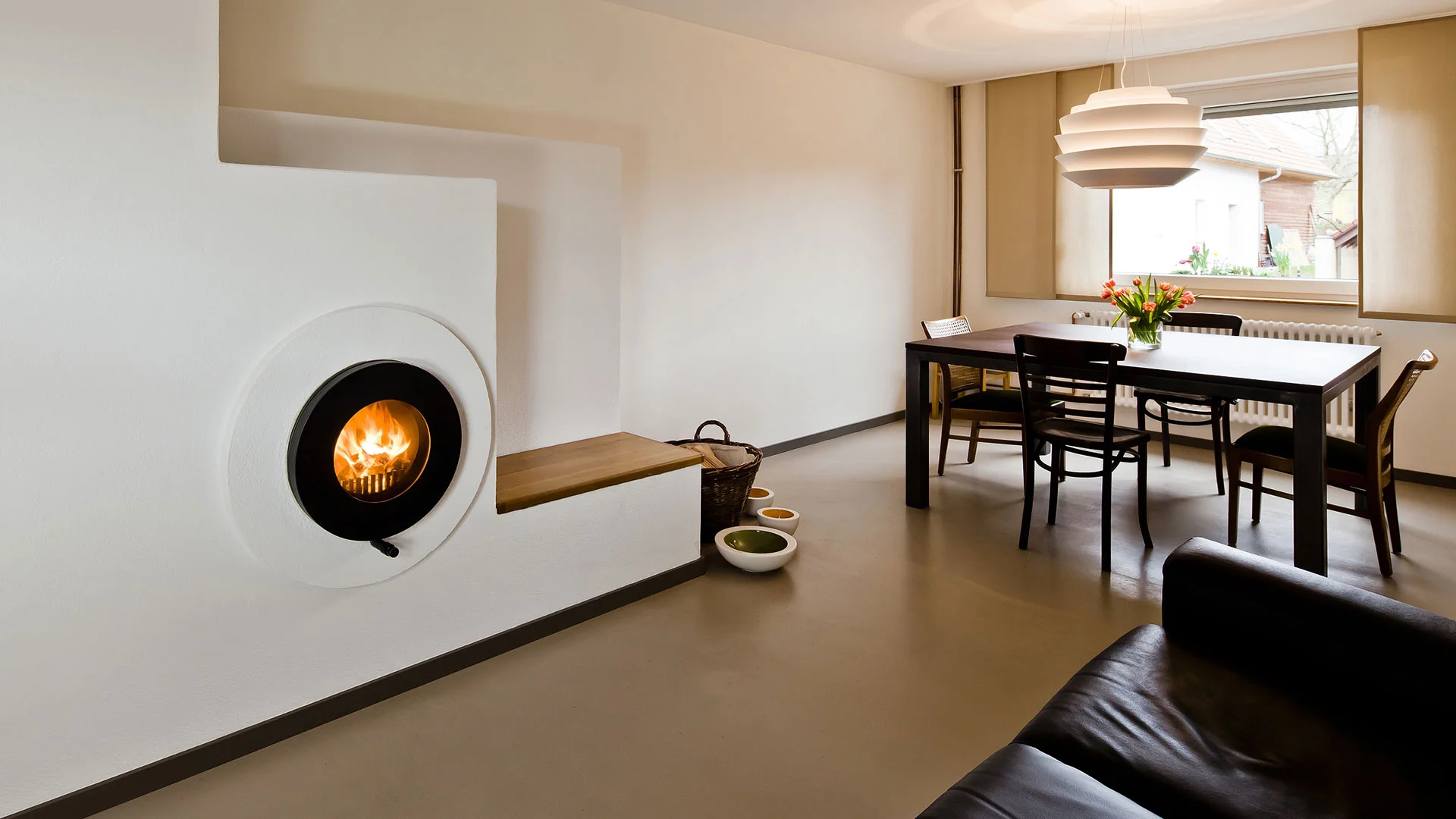Schwarzer firetube Ofen mit sichtbarem Feuer, in eine weiße Wand integriert; im Hintergrund ein Esstisch.