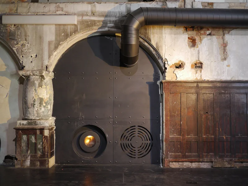 In eine rustikale Wand mit Türrahmen integrierter firetube Ofen aus dunklem Metall mit sichtbarem Feuer; unten links die Brennkammer, unten rechts eine Belüftungsöffnung.