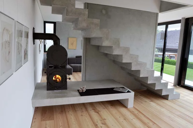 Schwarzer firetube Ofen mit sichtbarem Feuer und Garkammer über der Brennkammer, wandmontiert in modernem Wohnzimmer mit integrierter Sitzgelegenheit; daneben eine rustikale Betontreppe und Fenster im Hintergrund.