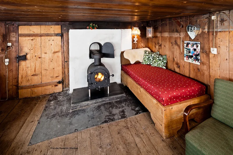 Gemütliches Zimmer mit Holzboden und Holzwänden, kleines Bett mit rotem Überzug; in der Mitte schwarzer firetube Ofen mit sichtbarem Feuer.