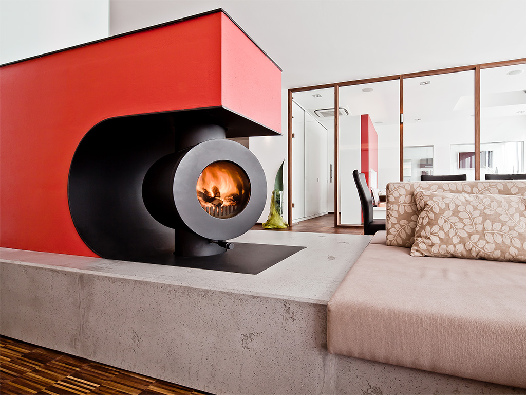 In eine Wohnlandschaft integrierter schwarzer firetube Speicherofen mit sichtbarem Feuer in rotem Wandausschnitt mit Sitzfläche; im Hintergrund ein Raum mit Glaswänden.